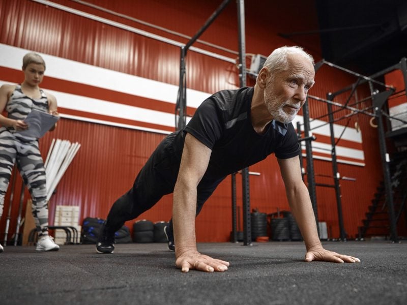 Confident sporty sixty year old man with beard doing push ups wearing stylish black sports clothes while his coach with clipboard writing down his results. Age, retirement, health and vitality